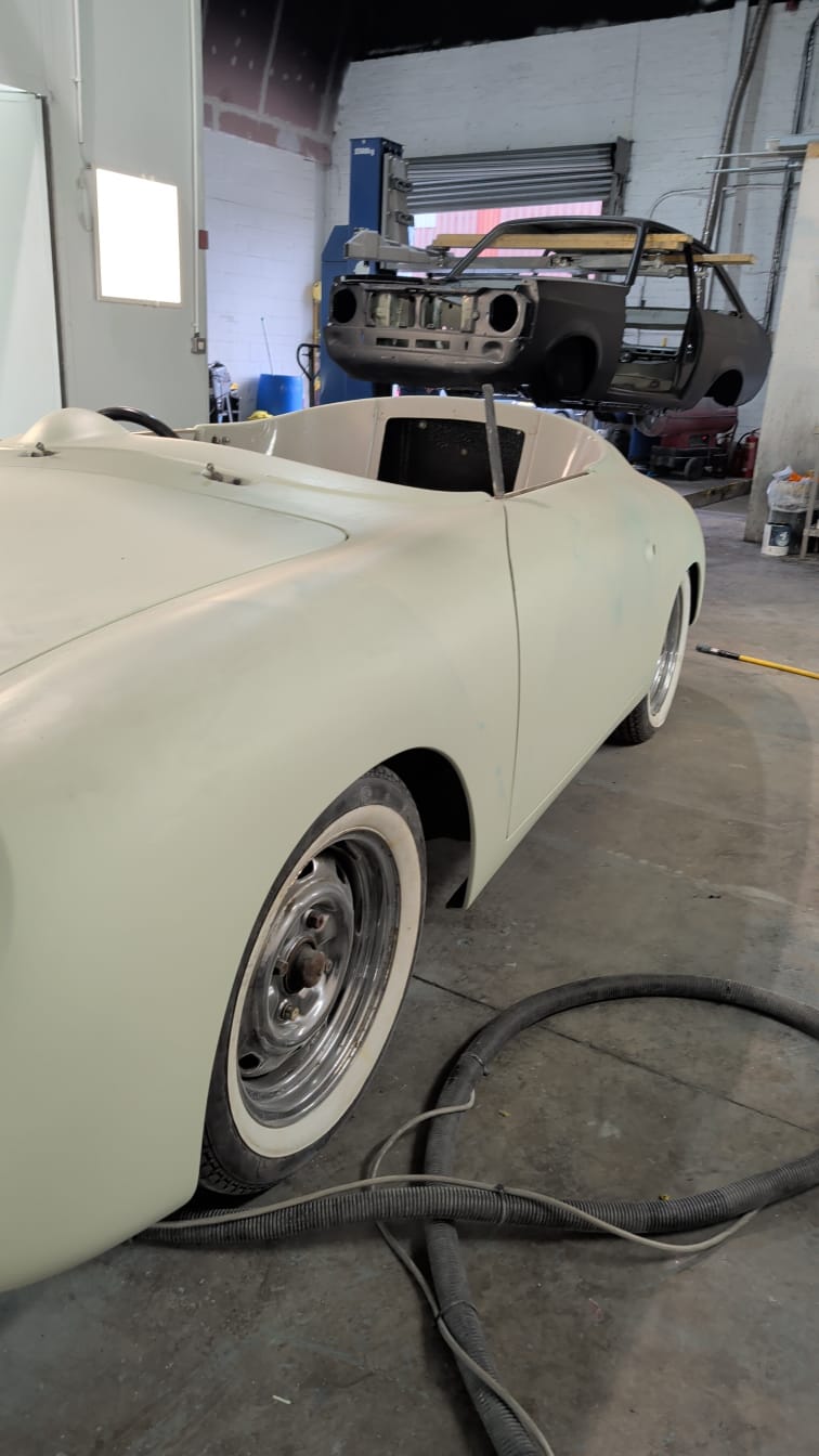 A classic sports car in primer on the workshop floor, with a bare shell up on the lift behind it